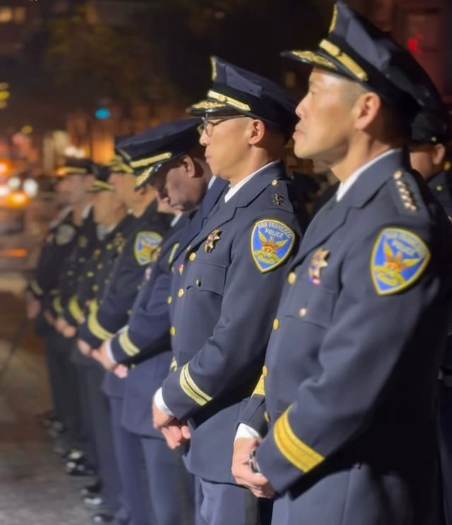 A line of San Francisco police officers in uniform stand solemnly outdoors at night, wearing peaked caps and gold-trimmed jackets.